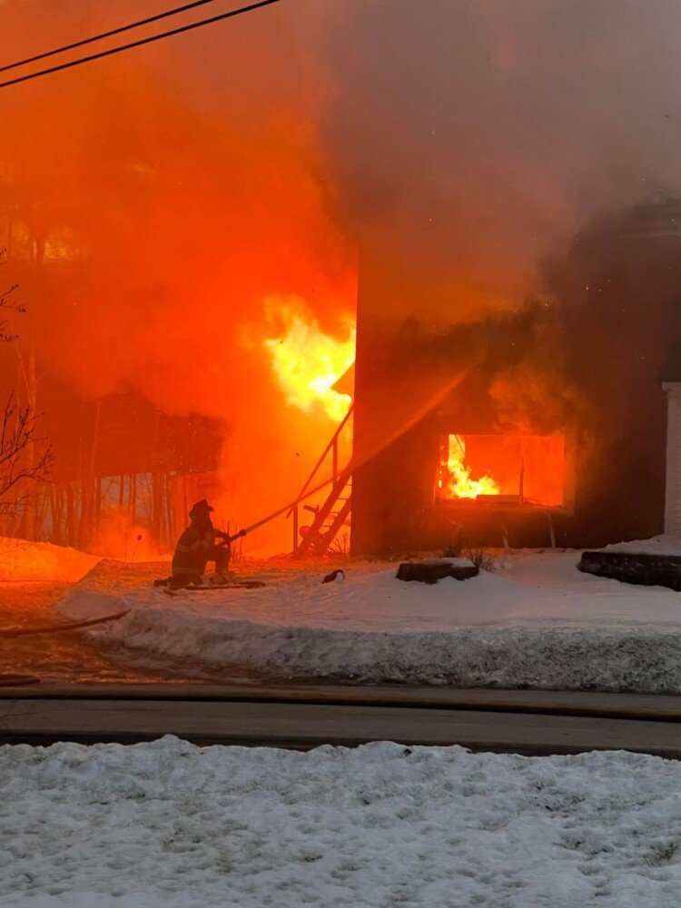 A firefighter kneels in the snow Saturday night as he douses a blaze at a century-old duplex home on Tallwood Drive in Readfield.
