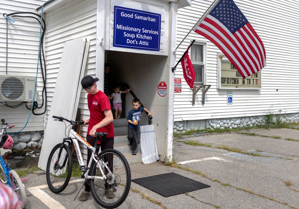 A boy stands at a bike in front of a building that two other young kids are exiting down a set of stairs.