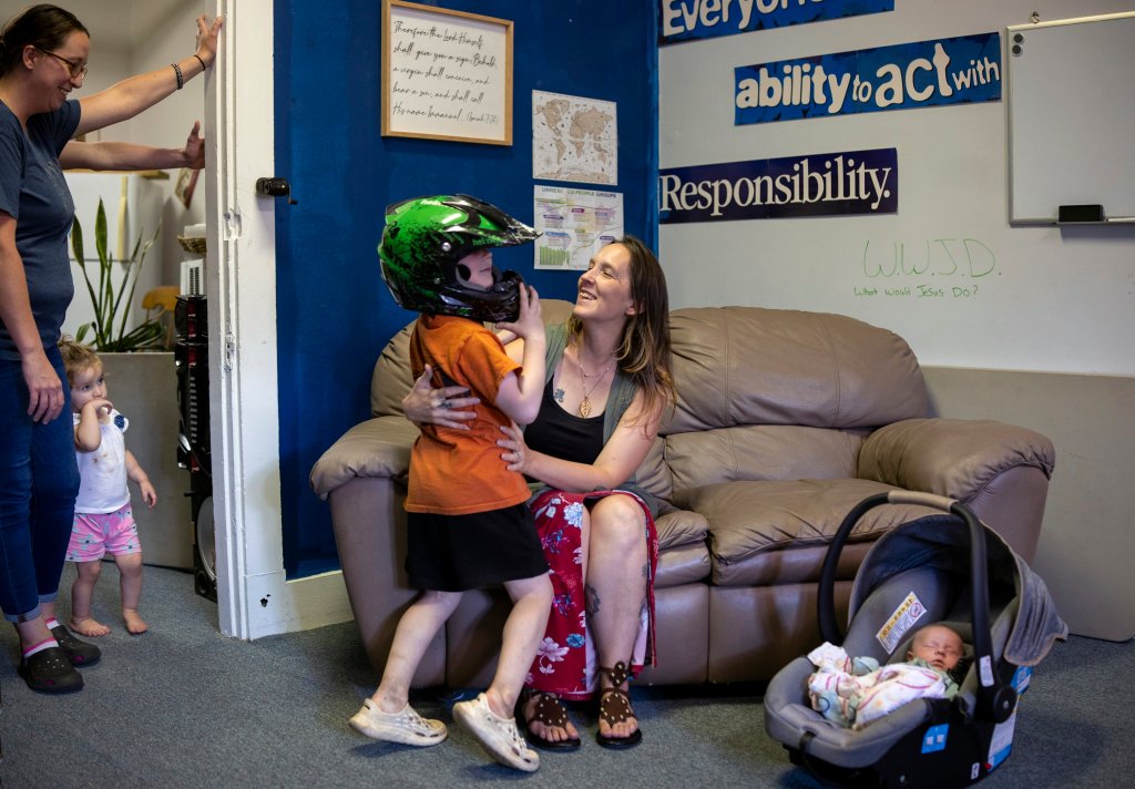 A woman looks at a young boy in a green helmet. A sleeping baby in a carseat sits next to them, and another woman and toddler look on.